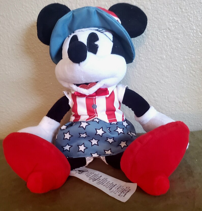 May include: A Minnie Mouse plush toy wearing a patriotic outfit. The toy has a blue hat, a red, white, and blue striped top, and a blue skirt with white stars. The plush has large red shoes and black ears.