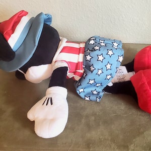 May include: A plush toy in the shape of a cartoon character, wearing patriotic attire. The hat is red, white, and blue. The shirt has red and white stripes, and the shorts are blue with white stars. The character has black fur, white gloves, and red shoes.