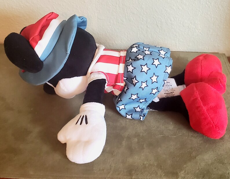 May include: A plush toy in the shape of a cartoon character, wearing patriotic attire. The hat is red, white, and blue. The shirt has red and white stripes, and the shorts are blue with white stars. The character has black fur, white gloves, and red shoes.