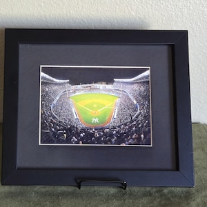Puede incluir: Una fotografía enmarcada de un estadio de béisbol de noche, con un campo verde y una multitud de personas en las gradas. El estadio está iluminado con luces, y el cielo está oscuro. La foto está enmarcada en un marco negro con un paspartú negro.