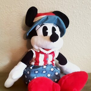 May include: A plush Minnie Mouse doll wearing a patriotic outfit. She has a red, white, and blue hat, a red and white striped top, and a blue skirt with white stars. Her large red shoes are visible.