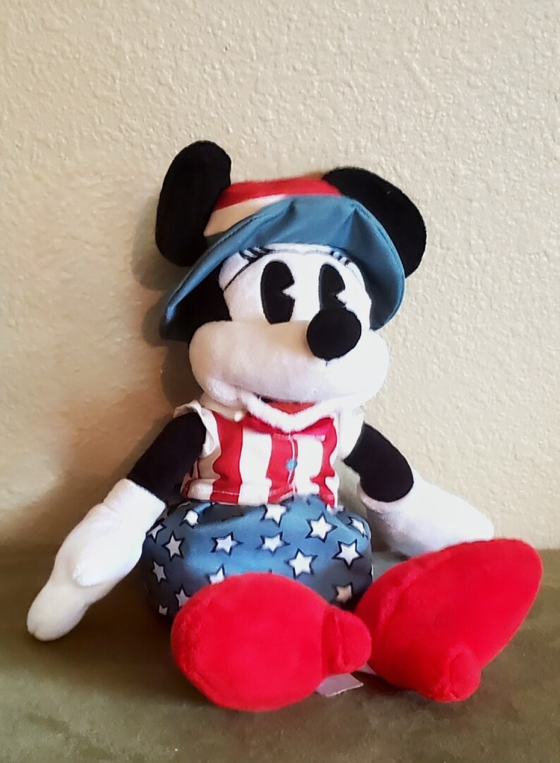 May include: A plush Minnie Mouse doll wearing a patriotic outfit. She has a red, white, and blue hat, a red and white striped top, and a blue skirt with white stars. Her large red shoes are visible.