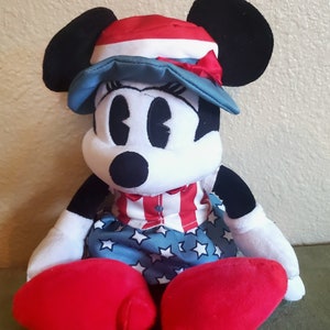 May include: A plush Minnie Mouse doll wearing a patriotic outfit. The doll has a red, white, and blue hat and dress. Her large red shoes and black ears are prominent. The doll is a classic Disney character.