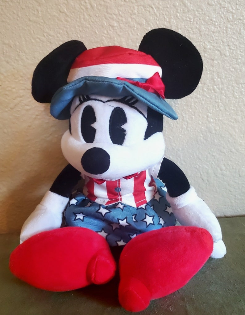 May include: A plush Minnie Mouse doll wearing a patriotic outfit. The doll has a red, white, and blue hat and dress. Her large red shoes and black ears are prominent. The doll is a classic Disney character.