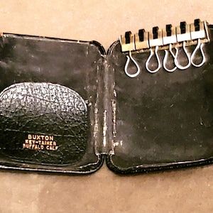 Może przedstawiać: Otwarty, czarny skórzany futerał na klucze ze złotym metalowym uchwytem na klucze. Na etui napis "BUXTON KEY-TAINER BUFFALO CALF". Uchwyt na klucze ma kilka kółek na klucze. Etui jest otwarte, ukazując wnętrze.