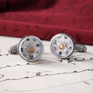 Könnte beinhalten: Ein Paar silberne Kompass-Manschettenknöpfe mit weißem Zifferblatt und schwarzen Markierungen. Die Kompassnadeln zeigen nach Norden.