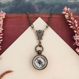 Puede incluir: Un delicado collar de oro con un colgante de brújula. La brújula tiene una cara blanca con marcas negras y una aguja azul. El collar está sobre una superficie blanca con flores rosas.