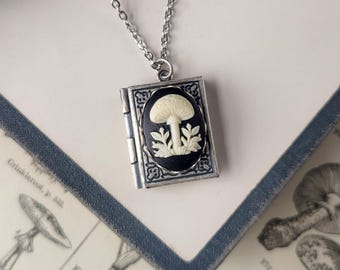 Puede incluir: Un collar plateado con un colgante en forma de libro. El colgante presenta un camafeo ovalado negro con un diseño de hongo blanco. El collar descansa sobre una ilustración botánica y un libro azul.