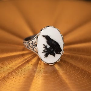 May include: A silver ring with an oval, white face featuring a black raven perched on a branch. The ring has an ornate, filigree design on the band. The raven is detailed, and the ring is set against a gold background.