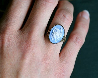 May include: A ring with a blue oval cameo featuring a white flower design. The ring is worn on a person's finger.