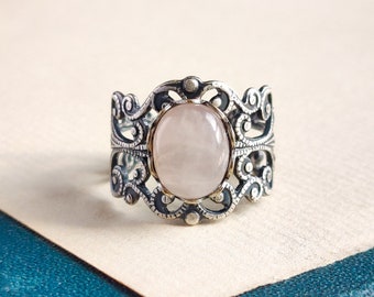 May include: A silver ring with an oval-shaped, light pink gemstone set in a filigree design.