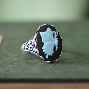 Peut inclure: Bague en argent avec un camée noir ovale représentant un papillon bleu clair. La bague a un anneau orné de filigrane. Le papillon est de profil, les ailes déployées. La bague est posée sur un fond vert.