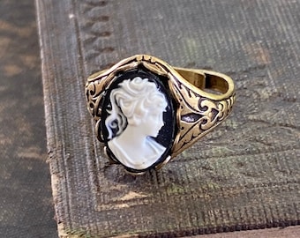 May include: A gold ring with a black and white cameo featuring a woman's profile. The ring has a detailed floral design.