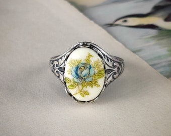 May include: Silver-toned ring featuring an oval, cream-colored centerpiece with a blue rose design. The ring has an ornate, filigree-style band. The rose has blue petals and green leaves, with gold accents. A vintage-inspired piece.