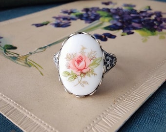 Könnte beinhalten: Ein silberner Ring mit einem weißen Porzellan-Kamee, der eine rosa Rose mit grünen Blättern zeigt.