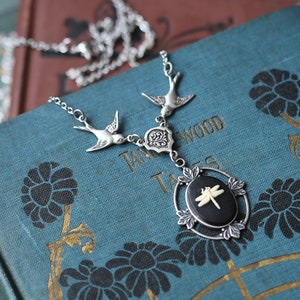 May include: A silver necklace with a dragonfly pendant. The pendant is black with a white dragonfly and is set in a silver frame. The necklace is on top of a stack of old books with a blue cover.