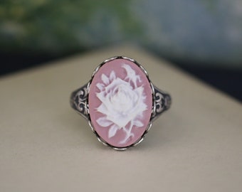 Könnte beinhalten: Ein silberner Ring mit einem rosafarbenen Kamee, der eine weiße geschnitzte Rose zeigt.