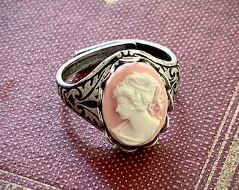 May include: A silver ring with a pink cameo featuring a woman's profile. The cameo is set in a detailed floral design.