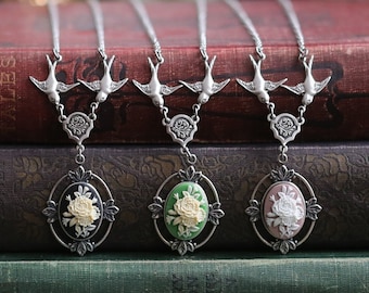 May include: Three silver necklaces with delicate bird charms and oval cameo pendants featuring white flowers on a green, black, and pink background. The necklaces are displayed on a stack of books.