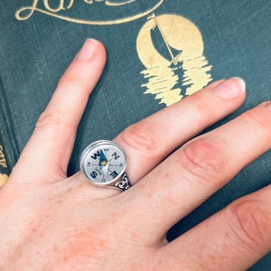 May include: A hand wearing a silver ring with a compass face. The ring is on a finger of a hand resting on a green book with gold lettering that reads "Lake Beautiful" and "William A. Quayle".