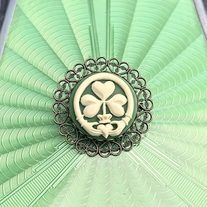 Puede incluir: Broche de camafeo redondo, verde y blanco, con diseño de trébol, engastado en un marco de filigrana ornamentado, color bronce. El broche está centrado en un fondo verde claro en forma de abanico con líneas grabadas.