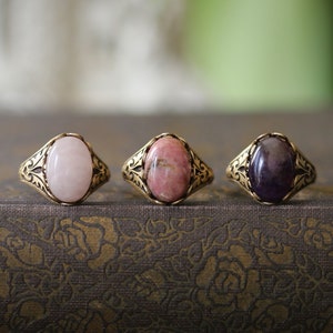 May include: Three gemstone rings with oval-shaped stones in pink, reddish-pink, and purple hues. The rings have ornate, gold-colored bands with decorative patterns. The rings are displayed on a textured, dark brown surface.