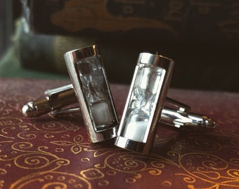 May include: A pair of silver cufflinks with an hourglass design. Each cufflink features a rectangular frame with a clear center, showcasing a miniature hourglass filled with white sand. The cufflinks rest on a burgundy leather-bound book.