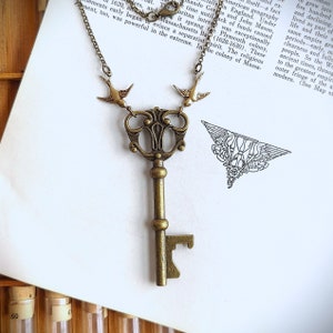 May include: Antique brass key pendant necklace with two bird charms. The key has an ornate design and is suspended from a delicate chain. The necklace is displayed on a vintage book page.