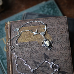 May include: A silver chain necklace with a cameo pendant featuring a white angel with wings. The pendant is surrounded by a black oval frame with silver accents. The necklace is laying on a vintage book with the words "A Face Illumine" on the cover.