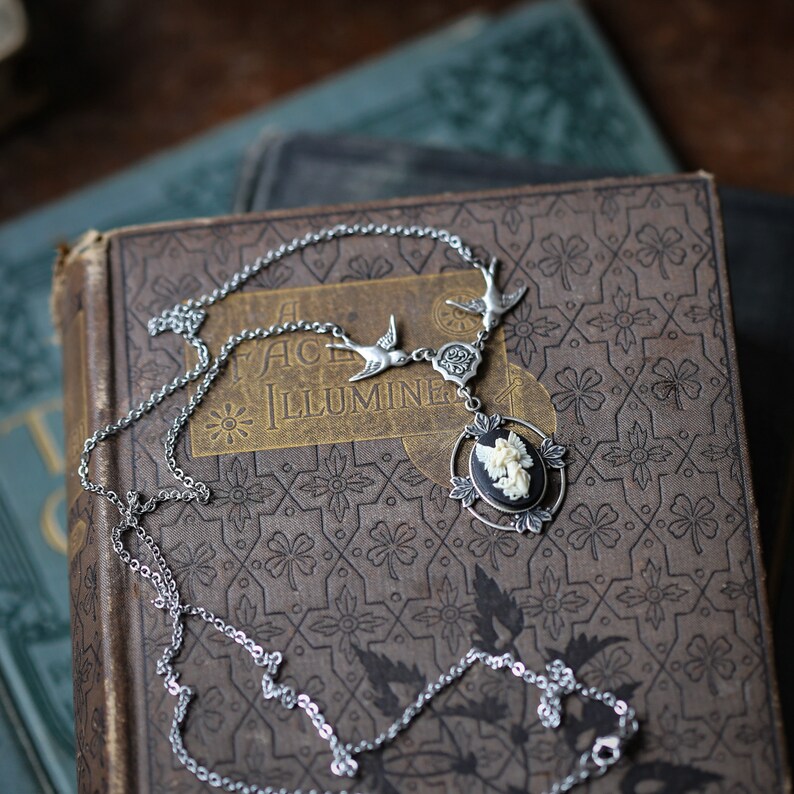 May include: A silver chain necklace with a cameo pendant featuring a white angel with wings. The pendant is surrounded by a black oval frame with silver accents. The necklace is laying on a vintage book with the words "A Face Illumine" on the cover.