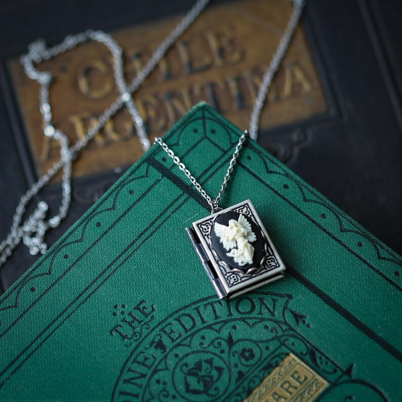 May include: A silver chain necklace with a locket pendant featuring a white cameo of an angel. The locket is open and the chain is draped over a green book with gold lettering that reads "The Illustrated Edition".