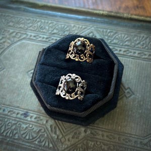 May include: Two rings with black gemstones set in ornate silver and gold filigree. The rings are displayed on a black velvet cushion.