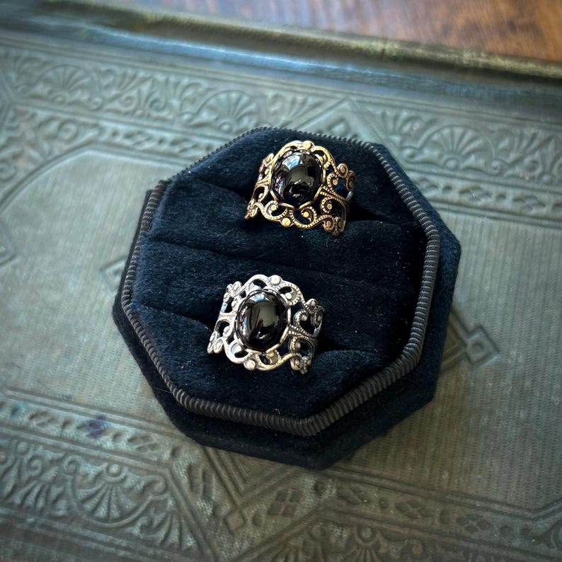 May include: Two rings with black gemstones set in ornate silver and gold filigree. The rings are displayed on a black velvet cushion.