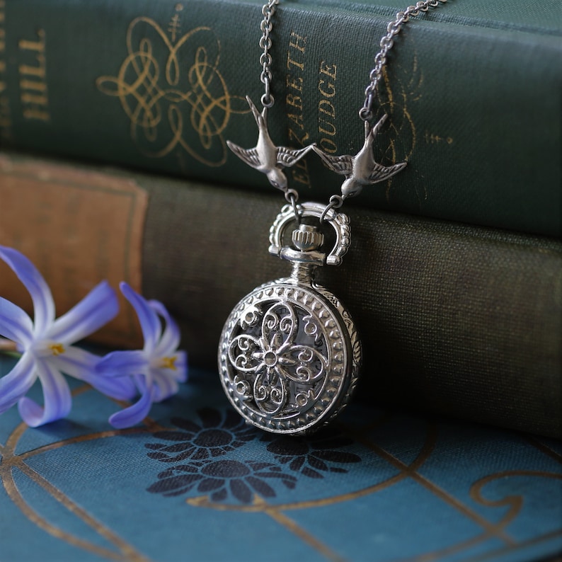 May include: A silver pocket watch with a delicate floral design hangs from a chain with two silver bird charms. The watch is resting on a stack of vintage books with a blue cover.