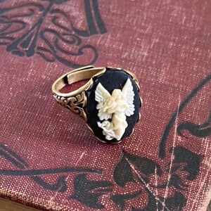 May include: A gold ring with a black oval cameo featuring a white carved angel with wings. The ring has a detailed filigree design.