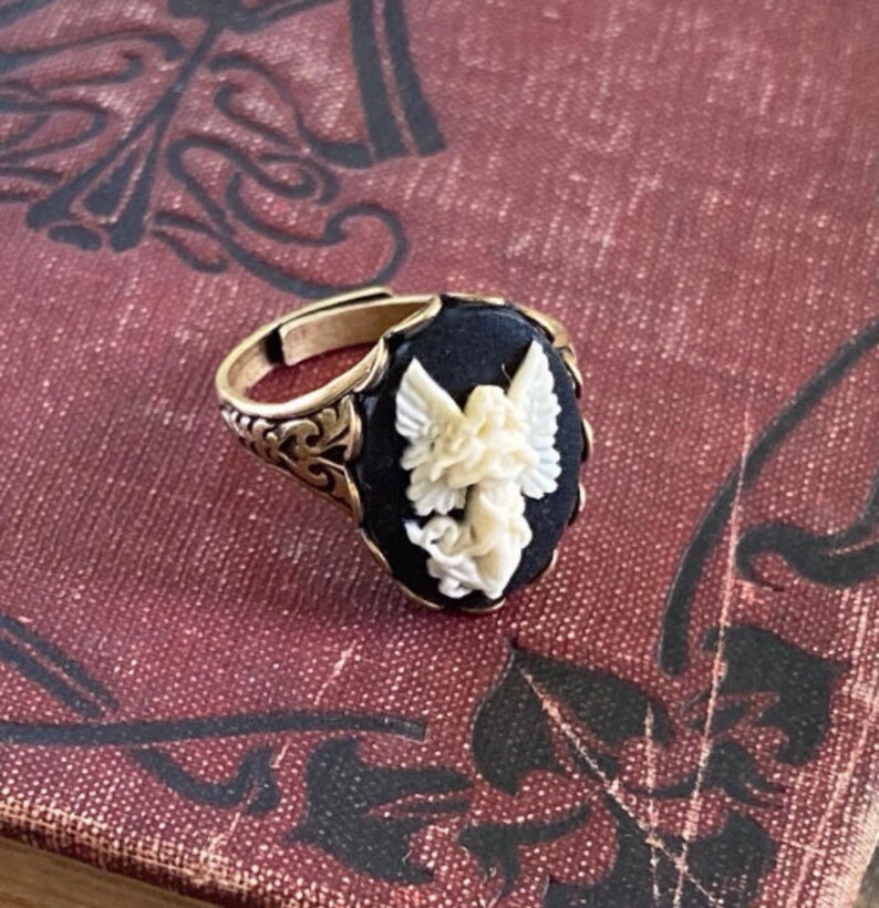 May include: A gold ring with a black oval cameo featuring a white carved angel with wings. The ring has a detailed filigree design.