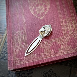 Puede incluir: Un marcap&aacute;ginas de metal plateado con un intrincado dise&ntilde;o de mecanismo de reloj, colocado sobre un libro rojo vintage. El dise&ntilde;o del marcap&aacute;ginas incorpora engranajes y otros elementos mec&aacute;nicos, a&ntilde;adiendo una est&eacute;tica steampunk. El libro tiene patrones dorados en relieve.