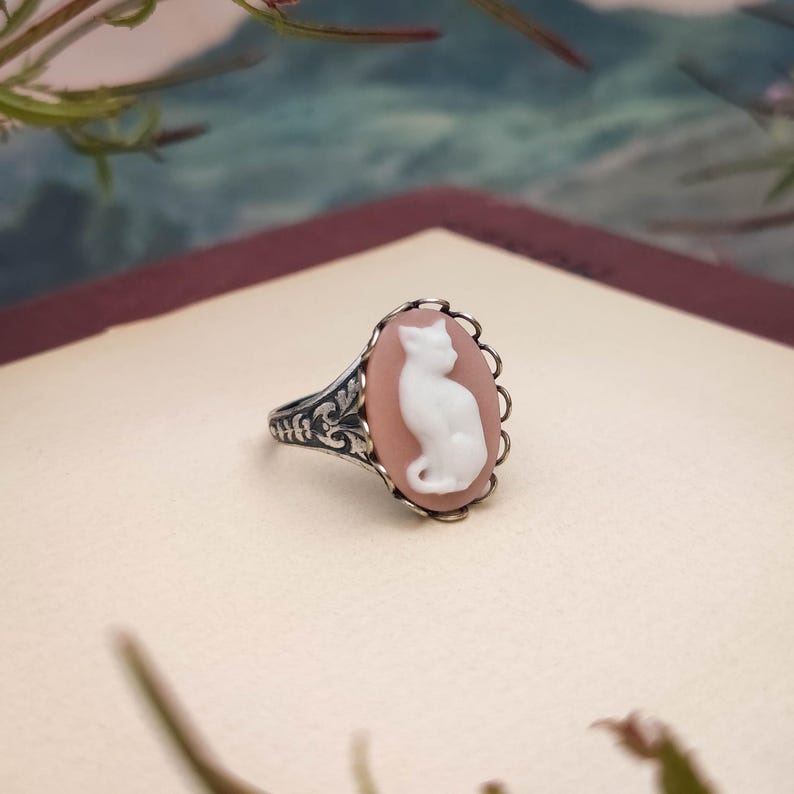 May include: A silver ring featuring an oval cameo with a white cat silhouette set against a soft peach-colored background. The metal band displays intricate engraved floral patterns along the sides, and the cameo is framed by a scalloped edge.