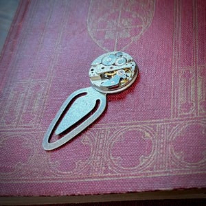 Puede incluir: Un marcap&aacute;ginas de metal plateado con un dise&ntilde;o de mecanismo de reloj. El marcap&aacute;ginas presenta un cuerpo en forma de l&aacute;grima con una esfera de reloj circular en la parte superior, que muestra engranajes y componentes intrincados. Descansa sobre un libro burdeos texturizado.