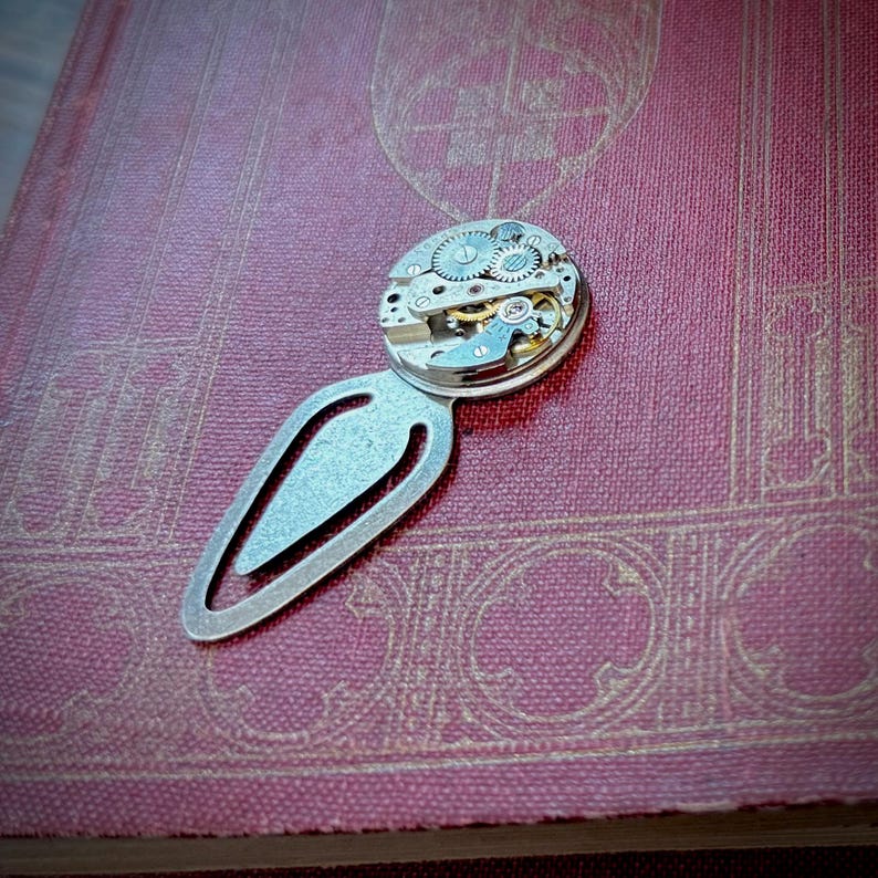 Puede incluir: Un marcap&aacute;ginas de metal plateado con un dise&ntilde;o de mecanismo de reloj. El marcap&aacute;ginas presenta un cuerpo en forma de l&aacute;grima con una esfera de reloj circular en la parte superior, que muestra engranajes y componentes intrincados. Descansa sobre un libro burdeos texturizado.