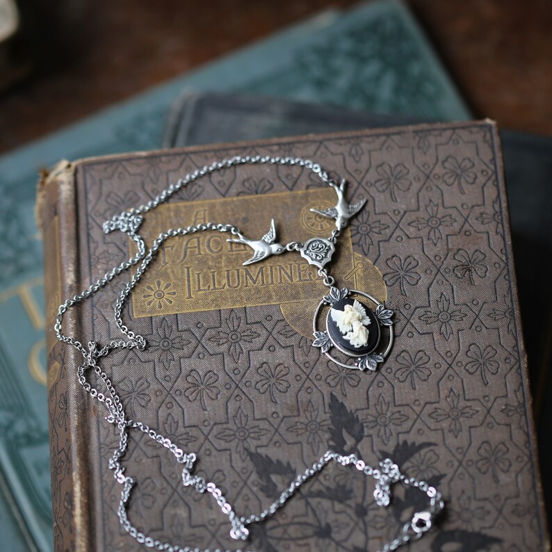 May include: A silver chain necklace with a cameo pendant featuring a white angel. The pendant is surrounded by a black oval and three silver birds. The necklace is lying on a vintage book with the text "A Face Illumine".