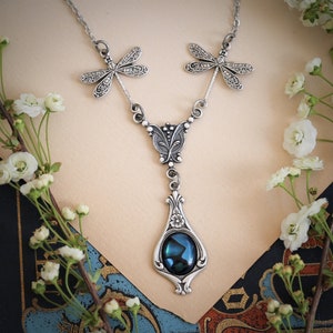 May include: A silver necklace with a blue abalone shell pendant and dragonfly accents. The necklace is on a white background.