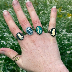 May include: A hand wearing four rings with cameo designs. The rings feature a white rabbit, a white cat, a mermaid, and a black cat. The rings are on a finger with a gold snake ring.