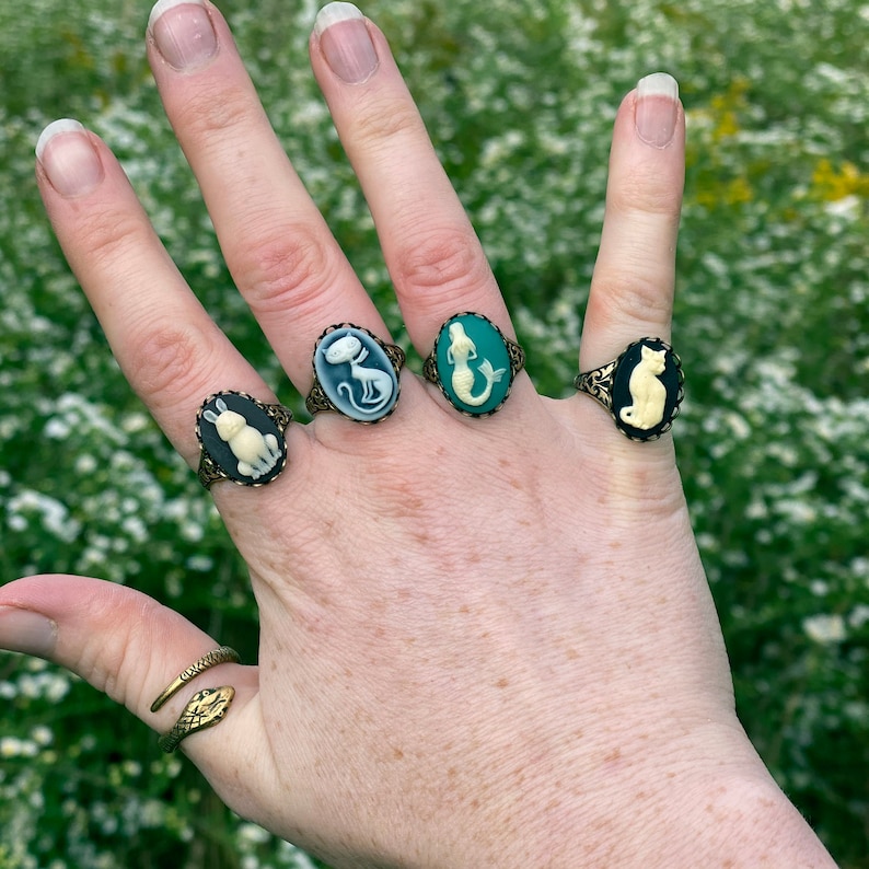 May include: A hand wearing four rings with cameo designs. The rings feature a white rabbit, a white cat, a mermaid, and a black cat. The rings are on a finger with a gold snake ring.