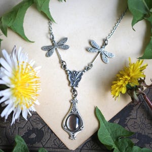 May include: A silver-toned necklace featuring a dragonfly design. The necklace has two dragonfly pendants at the top, connected to a central pendant with a teardrop-shaped stone. The necklace is surrounded by green leaves and yellow flowers.