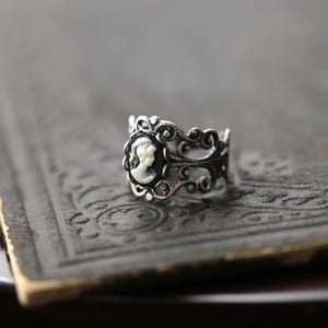 Pode incluir: Um anel de prata com um camafeu preto e branco com o perfil de uma mulher. O anel tem um design delicado e ornamentado.