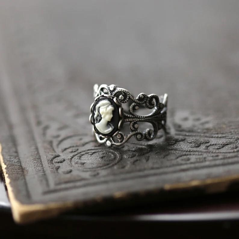 Pode incluir: Um anel de prata com um camafeu preto e branco com o perfil de uma mulher. O anel tem um design delicado e ornamentado.