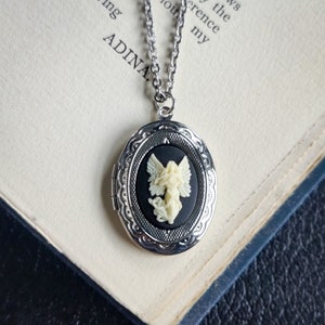 May include: A silver locket necklace with a black cameo featuring a white angel with wings. The locket is oval shaped and has a detailed design.