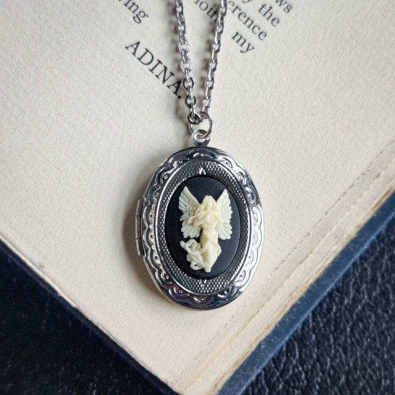 May include: A silver locket necklace with a black cameo featuring a white angel with wings. The locket is oval shaped and has a detailed design.