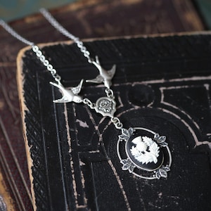 May include: A silver necklace with a black cameo featuring a white thistle. The necklace has two silver bird charms and a silver filigree charm.
